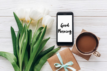 Obraz na płótnie Canvas good morning text sign on phone screen and stylish gift and tulips and coffee on white wooden rustic background. flat lay with flowers and gadget, space for text. spring concept