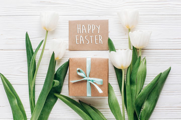 happy easter on greeting card with stylish present box and tulips on white wooden rustic background. flat lay mock up with flowers and empty paper with space for text.