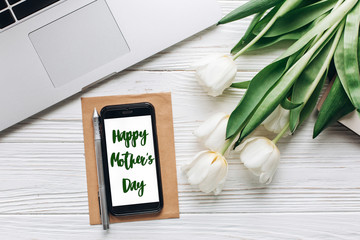 happy mothers day text sign on phone screen and laptop and tulips on white wooden rustic background. stylish flat lay with flowers and working gadgets with space for text. freelance. home working