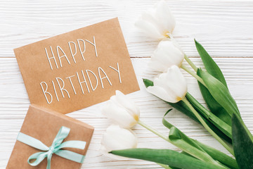 happy birthday text sign on stylish craft present with greeting card and tulips on white wooden rustic background. flat lay with flowers and gift with space for text.  greeting card