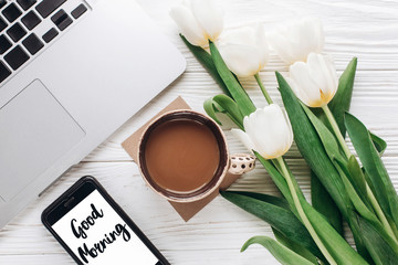 good morning text sign on phone screen and laptop with morning coffee and tulips on white wooden rustic background. stylish flat lay with flowers and working gadgets. freelance. home working