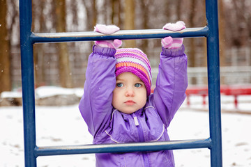 Portrait of little girl one year old playing outside in winter park playground. Toddler girl looking straight to the camera. Snowy weather.