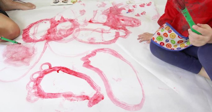 Three years blonde child with blue pants and mother with brush, in teamwork, painting  footprints with red watercolor on white paper in the floor
