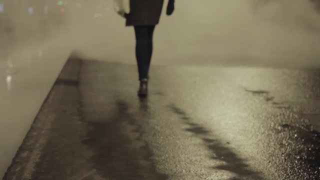 Girl in grey coat walk on night city street into white steam cloud, turn back