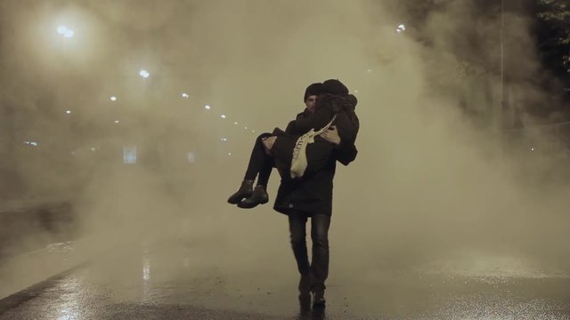 Man carry woman trough white steam cloud on night city street and kiss her