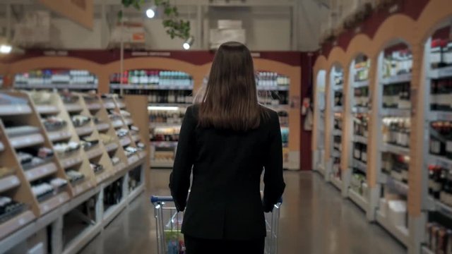 Woman shopping at the supermarket, follow shot from back of young woman with trolley in shopping center, alcohol store.