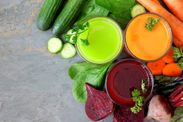 Drie glazen gezond groentesap met verspreide ingrediënten, boven uitzicht over een leisteen achtergrond