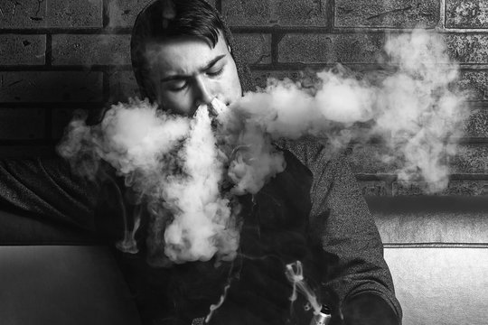 Young handsome white guy is admiting puffs of steam from the electronic cigarette. Vaping. Teenager listening to music. Black and white photo.