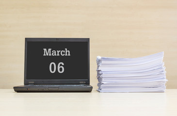 Closeup computer laptop with march 6 word on the center of screen in calendar concept and pile of work paper on wood desk and wood wall in work room textured background with copy space