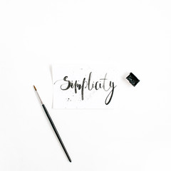 Minimalistic stylish composition with word Simplicity written in calligraphic style on paper with paint brush on white background. Flat lay, top view