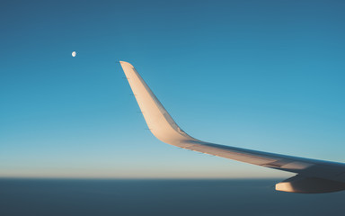 View from illuminator of dark teal morning misty horizon, clear sky during dawn without clouds, wing of modern airplane and partly closed moon
