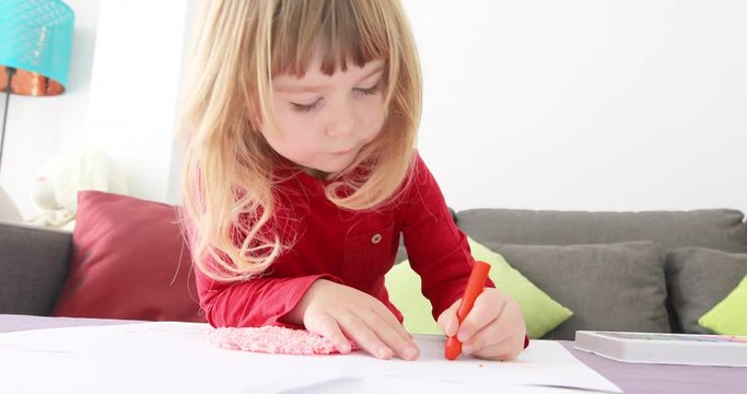 Three years blonde child red shirt with orange wax pencil o crayon in left hand painting white sheet paper on table