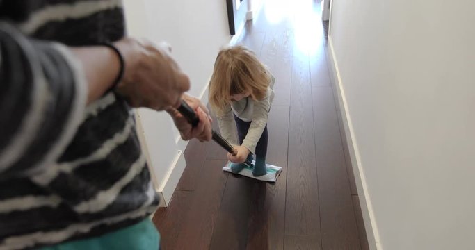 funny scene of three years old laughing child sliding on flat dust mop while woman sweeping the floor in corridor at home
