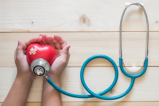 Young Child Kid Holding Red Love Heart Hand Exercise Ball With Bandage, MD Medical Doctor Physician's Stethoscope On Light White Wood Background: Nursing Children Day Care Healthcare Support Concept