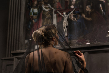 Fototapeta na wymiar young blond woman with beautiful hairstyle is covered by a black veil in black dress and precious jewelry kneeling at the altar in the background of the painting and the arch looks at Jesus and prays