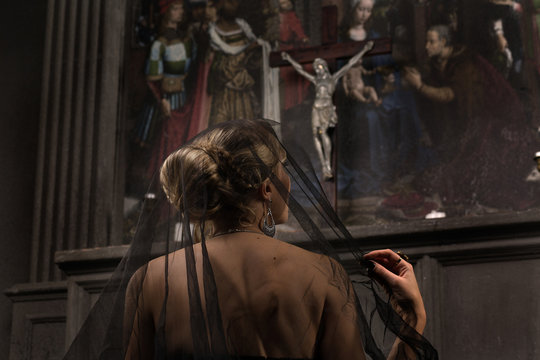 young blond woman with beautiful hairstyle is covered by a black veil in black dress and precious jewelry kneeling at the altar in the background of the painting and the arch looks at Jesus and prays