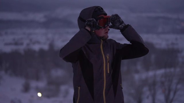 A man in a ski suit in the evening at the ski resort. Young man adjusts his goggles for skiing at night on the slopes. Winter sports keep muscles in a tone.