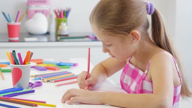 Little girl sitting at table drawing with colored pencils and showing her painting at camera.