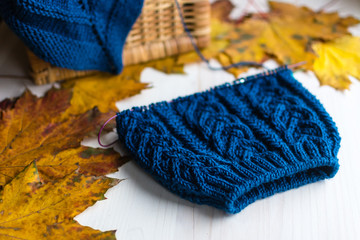 Knitted blue wool hat and needles on white table with dry maple leafs on background. Unfinished needlework in process.