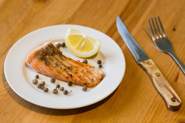 Portion of cooked salmon fillet with lemon slice on white plate with fork and knife on wooden table