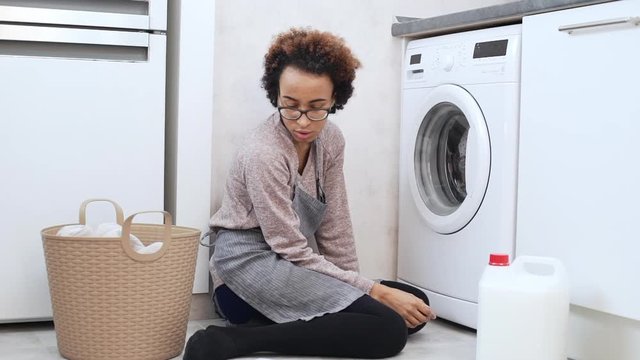 Young vexed african american female in glasses sitting on floor and trying to sort out how to use washing machine reading manual in slowmotion