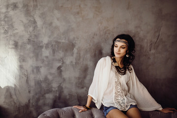 Charming lady in white ethnic shirt sits on grey sofa before the wall