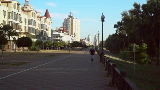 A young man is jogging ouside in the city across the path. The male is wearing trendy leggins and shorts outfit. He runs near the buildings along the path.