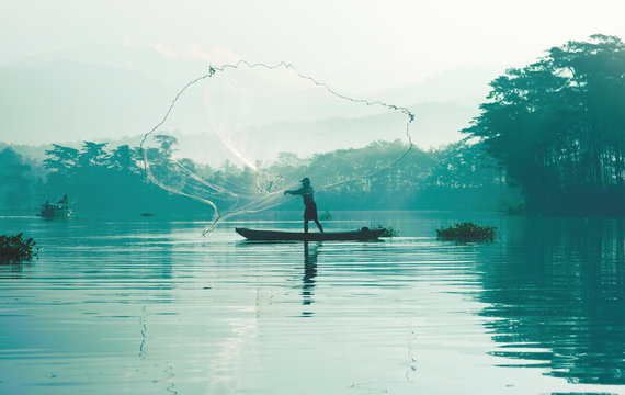 Fisherman casting out his fishing net in the river by throwing it high up into the air early in the blue colored morning to catch fish with his little fishing boat.