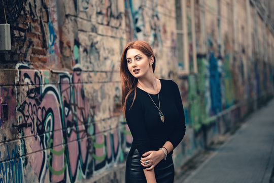Red-head cheeky young woman, giving inviting smile in front of street art colored wall. Arching on fleek eyebrow, having burgundy lips and smokey eyes makeup, creating naughty look.