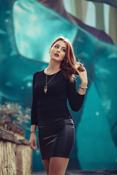 Red-head cheeky young woman, giving inviting smile in front of street art colored wall. Arching on fleek eyebrow, having burgundy lips and smokey eyes makeup, creating naughty look.