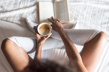 black woman reading a book and drinking coffee on bed with socks
