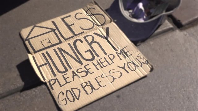 Homeless person on side of street has cardboard sign asking for help.
