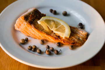 Portion of cooked salmon fillet with lemon slice on white plate with fork and knife on wooden table