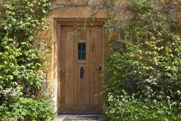 Fototapeta premium brown wooden doors in an old traditional English lime stone house with flowers, rosebush, climbing shrubs in the front garden, on summer day .