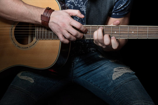 Man in torn jeans delicately playing an acoustic guitar, close-up, on a black background