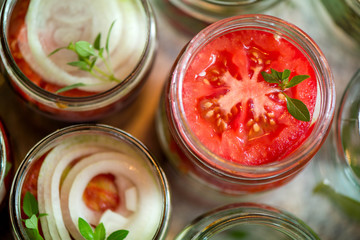 Canning fresh tomatoes with onions for winter in jelly marinade. A shot of basil leaves on top of a red ripe tomato slices and onion rings being put in jar.

