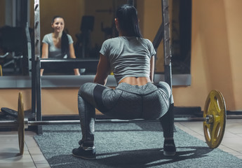 Slim girl bodybuilder lifting heavy barbell standing in front of the mirror while training in the...
