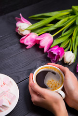 Bouquet of tender pink tulips and hands holding cup of coffee and marshmallows on black wooden background