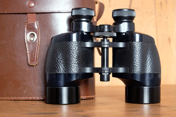 Vintage Porro prism black color binoculars and closed brown hard leather case on wooden background...