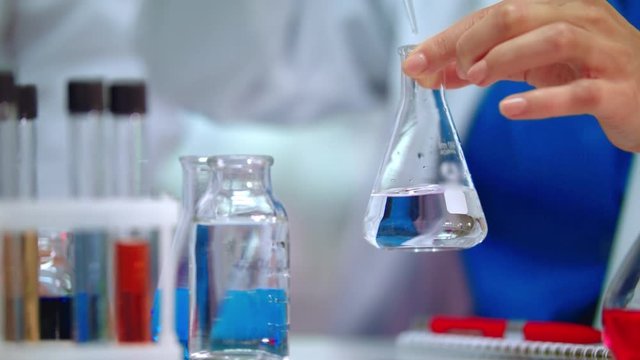 Chemical reaction in lab flask. Close up of female chemist hands doing chemical experiment in beaker. Chemist adding chemical reagent in pure liquid at conical flask. Chemical research