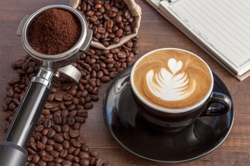 Coffee cup of latte art in the black color cup with some coffee beans, portafilter and notebook on wooden background