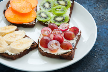 Variety slices of rye bread toast with fruits. Banana persimmon, kiwi, grape, sandwiches in a white plate on rusty background. Flat lay.