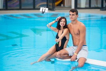 Young pair - man and woman sitting by the pool with perfect aqua water and making selfie photo on the phone with monopod on the luxury resort