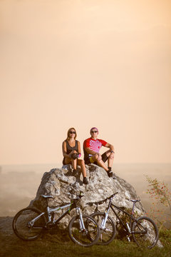 Cyclists are sitting on a large stone on the precipice of a cliff next to them are sports bikes at summer evening. Blurred background