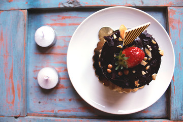 Chocolate cake with strawberries on top, sprinkled with nuts on a white plate. candles and green plants in the background.