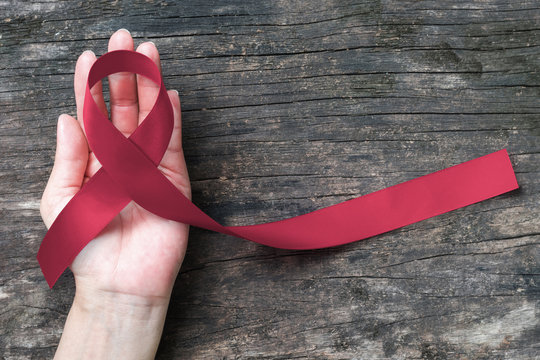 Burgundy ribbon for multiple myeloma cancer awareness on human hand old aged wood background: Satin fabric symbolic concept raising public support campaign on people living w/ disease