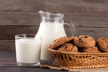 jug and glass of milk with oatmeal cookies in a wicker basket on a wooden background