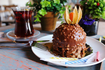 Chocolate cake with a berry on top, sprinkled with nuts on a white plate.with turkish tea and  candles and green plants in the background.