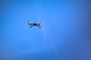 Black, small, mini quadrocopter flying high in the air, taking photos, recording footage from above. Flying drone with four motors, yellow propellers, camera on clear blue sky background. Surveillance