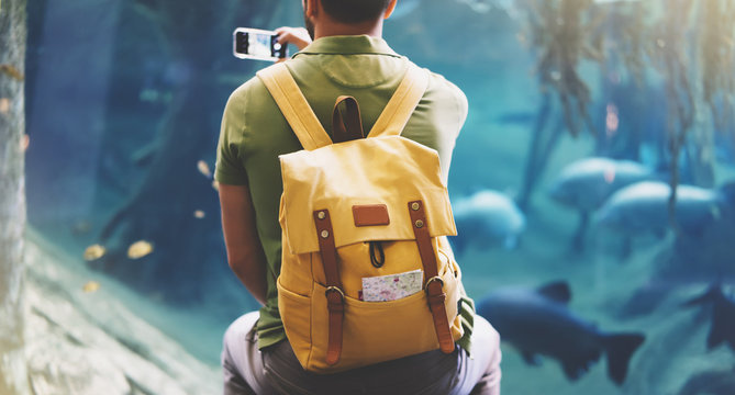 Hipster with yellow backpack, map, phone. View from back tourist traveler on background blue sea aquarium. Person hiker in oceanarium museum Barcelona and photographer pictures fish smartphone mockup